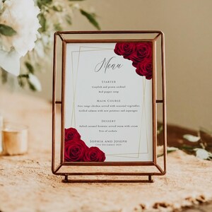 May include: A framed menu card with a copper-colored metal frame. The menu features a white background with gold geometric lines and red roses. The menu text includes the word "Menu" and the names "Sophia and Joseph".