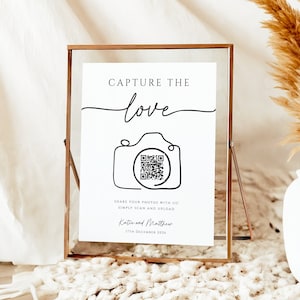 May include: A white sign with a gold frame that says "Capture the love" with a line drawing of a camera with a QR code inside. The sign is on a white rug with a vase of dried pampas grass.