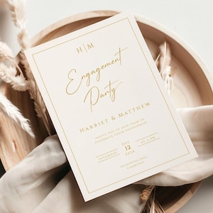 Könnte beinhalten: Eine cremefarbene Einladungskarte zur Verlobungsfeier mit goldenem Text. Die Einladung mit den Worten "Engagement Party" liegt auf einem Holztablett mit getrockneten Pflanzen und einem cremefarbenen Tuch. Datum und Uhrzeit sind ebenfalls angegeben.