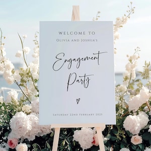 May include: A white sign on a wooden easel with the words "Welcome to Olivia and Joshua's Engagement Party" in black script. The sign is surrounded by white and pink flowers, with a blurred ocean background.