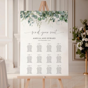 May include: A white wedding seating chart with a green eucalyptus and floral design. The chart has the text "find your seat" and "Amelia and Edward" with the date "4th September 2020". The chart is displayed on a wooden easel.