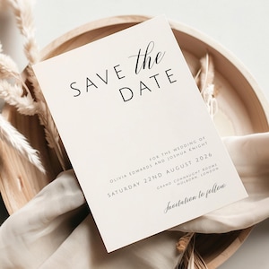 以下が含まれることがあります： ミニマルなデザインの「Save the Date」カード。カードは白地に黒文字で、「Save the Date」の文字が書かれており、「the」は筆記体になっています。カードには、Olivia Edwards と Joshua Knight の名前、日付は 2026 年 8 月 22 日土曜日、場所は Grand Connaught Rooms、Holborn London が記載されています。