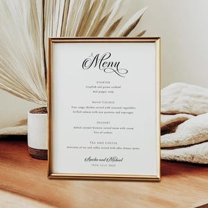 May include: A framed menu with a gold border, featuring elegant script text. The menu lists a starter, main course, dessert, and tea/coffee options. The menu is for Amelia and Michael, dated July 19th, 2025.