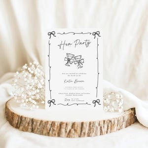 May include: A white invitation card with black text and illustrations, featuring the words "Hen Party" and two champagne glasses. The card includes details for Katie Brown and is decorated with bow accents. The card is displayed on a wooden slice.