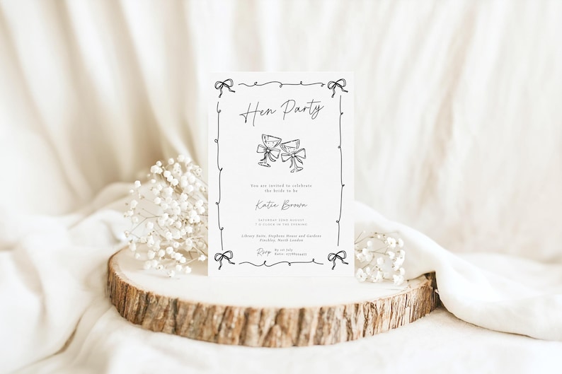 May include: A white invitation card with black text and illustrations, featuring the words "Hen Party" and two champagne glasses. The card includes details for Katie Brown and is decorated with bow accents. The card is displayed on a wooden slice.