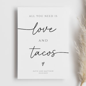 May include: White card with black text that reads "All you need is love and tacos" with a small heart symbol below the text. The text "Katie and Matthew 22.8.2023" is at the bottom of the card.