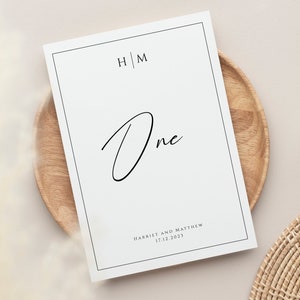May include: White wedding table number card with black border and text. The number "One" is written in a script font. The initials "H|M" and the names "Harriet and Matthew" and the date "17.12.2023" are printed in black.