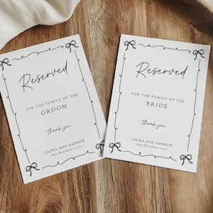 May include: Two white wedding place cards with black script that reads "Reserved" and "For the family of the Groom" and "For the family of the Bride". Each card has a decorative border with bows and the names Laura and Andrew.