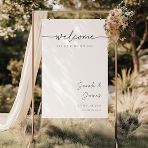 May include: A white wedding welcome sign with the words "welcome to our wedding" in elegant script. The sign also includes the names "Sarah & James", the date "17th June 2023", and "Gosfield Hall". The sign is decorated with flowers.
