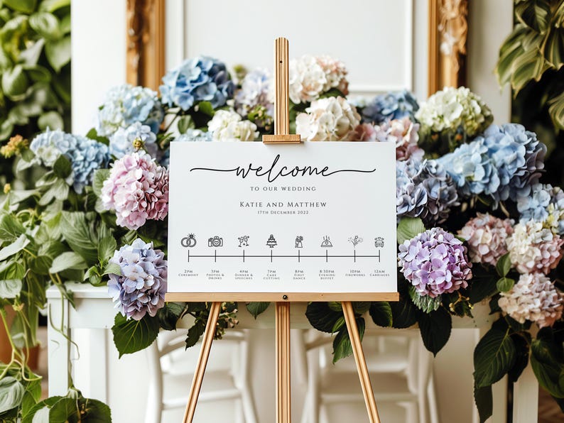 May include: A white wedding welcome sign with a black calligraphy script that reads "Welcome to our wedding Katie and Matthew 17th December 2022". The sign also includes a timeline of wedding events with icons for the ceremony, photos, dinner, cake cutting, and dancing.