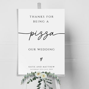 以下が含まれることがあります： 白い結婚式の看板に、黒い文字で「ピザになってくれてありがとう、私たちの結婚式」と書かれており、ハートマークと、ケイティとマシューの名前、そして2025年7月12日土曜日の日付が記されています。