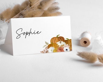 Puede incluir: Una tarjeta de sitio blanca con el nombre "Sophie" en escritura negra. La tarjeta presenta una ilustración de acuarela de calabazas y flores de otoño en tonos naranja, blanco y marrón. Una bobina de hilo blanco y flores secas están en el fondo.