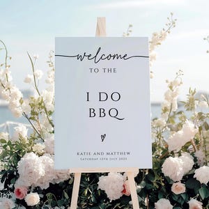 May include: White welcome sign with black calligraphy that reads "Welcome to the I Do BBQ Katie and Matthew Saturday 12th July 2025".
