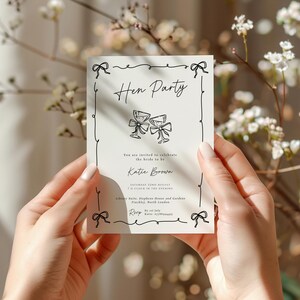 May include: A cream-colored invitation card with the words "Hen Party" in elegant script, featuring two champagne glasses. The card is held by hands and surrounded by floral elements. The invitation includes details for Katie Brown's celebration.
