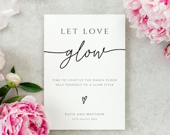 May include: White card with black text that reads "Let love glow" with a handwritten script font. The text continues with "Time to light up the dance floor. Help yourself to a glow stick." and a small heart symbol. The bottom of the card reads "Katie and Matthew, 27th August 2024".