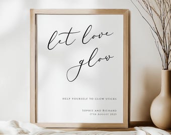 May include: A framed print with the words "let love glow" in elegant black script. Below, it reads "Help yourself to glow sticks" and the names "Sophie and Richard" with a date. The frame is light wood.