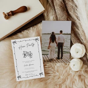 May include: A hen party invitation with black script and bow illustrations. A photo of a couple on a dock, white flowers, and a wooden stamp are also present. The items are arranged on a fluffy, light-colored surface.
