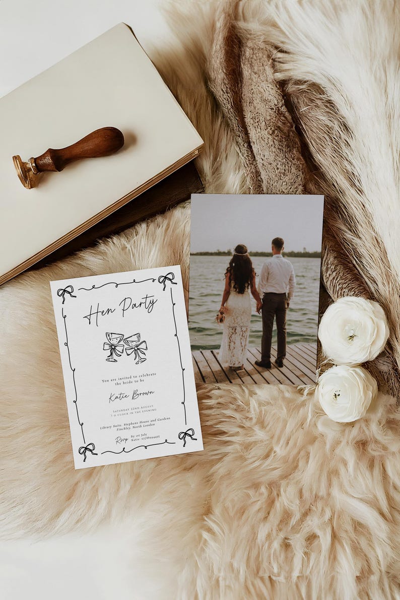 May include: A hen party invitation with black script and bow illustrations. A photo of a couple on a dock, white flowers, and a wooden stamp are also present. The items are arranged on a fluffy, light-colored surface.
