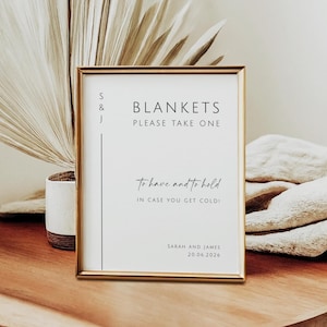 May include: A framed sign with gold trim, featuring the text "BLANKETS PLEASE TAKE ONE" in black. Below, it reads "to have and to hold, in case you get cold!" The sign includes names and a date. The sign is on a wooden surface.