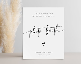 May include: White sign with black text that says "Grab a prop and remember to smile!" and "photo booth" with a heart symbol. Below the text is "Olivia and Joshua" and "27th August 2024".