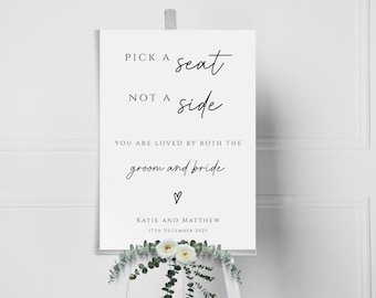 May include: White sign with black text that reads "Pick a seat, not a side. You are loved by both the groom and bride."  A heart symbol is below the text. The sign also includes the names "Katie and Matthew" and the date "17th December 2025".