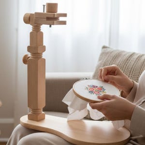 May include: A wooden embroidery stand with a light-colored finish. The stand features a curved base and adjustable arm to hold an embroidery hoop. The image shows a person working on a floral embroidery design.