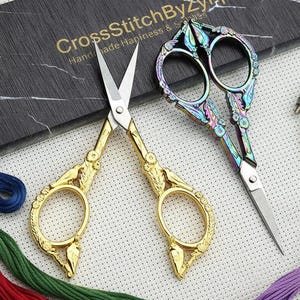May include: Two pairs of decorative embroidery scissors with ornate handles. One pair is gold-colored, the other has a rainbow iridescent finish. The scissors are next to a black marble-look box with the text "CrossStitchByZynp". Colorful embroidery floss and a wooden hoop are also visible.