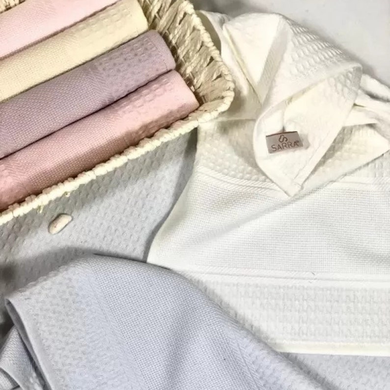 May include: A collection of waffle-weave towels in pastel shades of pink, cream, and lavender, neatly folded inside a woven basket. A white towel with a SARRA brand tag is displayed alongside a light blue towel, all featuring a textured pattern.