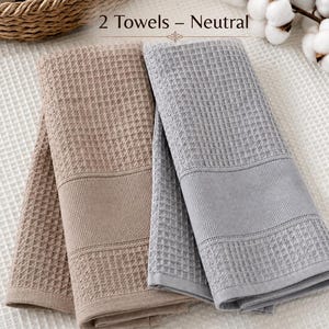 May include: Two folded waffle-weave towels, one in a light brown cappuccino color and one in a light gray color. The text 2 Towels - Neutral is centered above the towels, with the word Cappuccino below the brown towel and Gray below the gray towel.