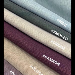 May include: Close-up of fabric swatches in various colors, including light blue, dark green, cream, and dark purple. The fabric has a textured appearance. The names of the colors are written on the fabric: #NILE, #SMOKED, #CREAM, #DAMSON, #OLD SACK, and #DARK LILAC.