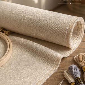 May include: A rolled-up, beige 16-count sparkle Aida fabric with a wooden embroidery hoop, embroidery floss, and gold scissors. The fabric has a subtle shimmer and is ideal for cross-stitch projects.