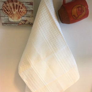 May include: A white kitchen towel featuring a textured waffle weave pattern with vertical striped bands. The towel hangs from a metal hook between a square wall plaque depicting a seashell and the word SHORE, and an orange ceramic mug.