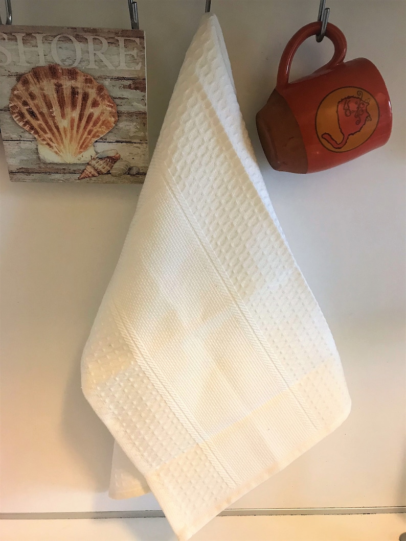 May include: A white kitchen towel featuring a textured waffle weave pattern with vertical striped bands. The towel hangs from a metal hook between a square wall plaque depicting a seashell and the word SHORE, and an orange ceramic mug.