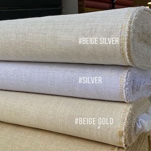 May include: A stack of fabric rolls in various neutral colors, including beige, silver, and dark beige. Each roll has a textured surface and a tag with a color description. The fabric is likely for sewing or crafting.