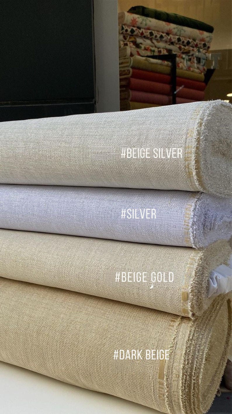 May include: A stack of fabric rolls in various neutral colors, including beige, silver, and dark beige. Each roll has a textured surface and a tag with a color description. The fabric is likely for sewing or crafting.