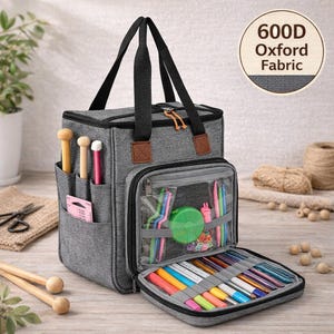May include: A gray knitting bag with black handles and brown accents. The bag has multiple pockets and compartments, including a clear zippered section and a lower compartment filled with colorful knitting supplies. The text "600D Oxford Fabric" is in a circle on the upper right.