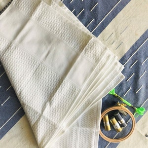 May include: A stack of white waffle weave towels folded in a fan shape on a blue and white striped fabric.  There are spools of embroidery floss and a wooden embroidery hoop in the lower right corner of the image.