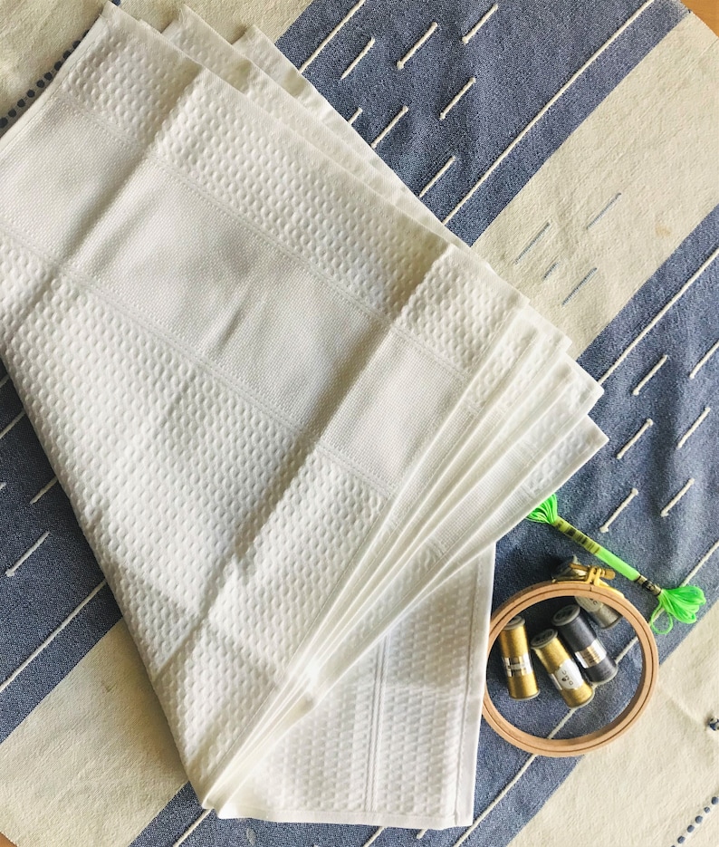 May include: A stack of white waffle weave towels folded in a fan shape on a blue and white striped fabric.  There are spools of embroidery floss and a wooden embroidery hoop in the lower right corner of the image.