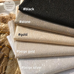May include: A stack of shimmering Aida fabric swatches in various colors, including black, stone, gold, beige gold, beige silver, and silver. The fabric has an opalescent shimmer effect and is 16 count. Text on the image reads "Sparkle Aida Fabric".