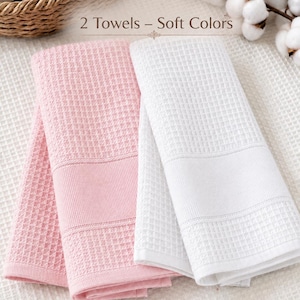 May include: Two folded waffle-weave towels, one in soft pink and one in white, featuring a textured grid pattern and a smooth horizontal band. The text "2 Towels &ndash; Soft Colors" is visible above, with "Pink" and "White" labels below each towel.