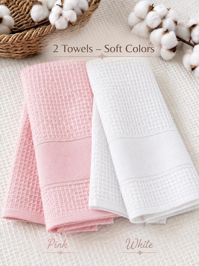 May include: Two folded waffle-weave towels, one in soft pink and one in white, featuring a textured grid pattern and a smooth horizontal band. The text "2 Towels &ndash; Soft Colors" is visible above, with "Pink" and "White" labels below each towel.