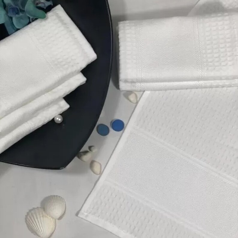 May include: A set of white towels with a waffle texture. The towels are folded and stacked on a black plate. There are two blue and white decorative stones on a white surface.