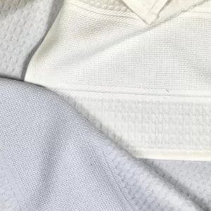 May include: A white waffle-weave fabric towel featuring a distinct textured grid pattern throughout the material. The textile displays a clean, uniform surface with a visible hemmed edge and a subtle, repeating square design across the entire piece.