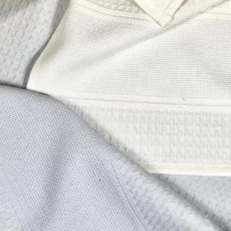 May include: A white waffle-weave fabric towel featuring a distinct textured grid pattern throughout the material. The textile displays a clean, uniform surface with a visible hemmed edge and a subtle, repeating square design across the entire piece.