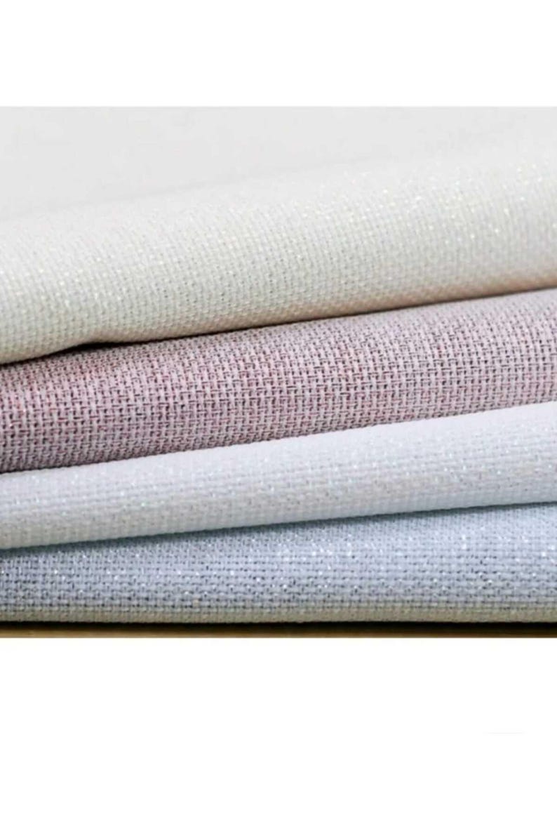 May include: Close-up of four fabric samples in varying shades of white, pink, and gray. The fabrics have a textured weave with a subtle shimmer, suggesting a decorative or upholstery material. The layers are neatly stacked.