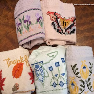 May include: Five folded fabric towels featuring cross-stitch embroidery. Designs include purple irises, a red and yellow butterfly, an orange leaf with the text "Thankful", a green geometric floral pattern, and yellow tulip-like flowers with green stems.