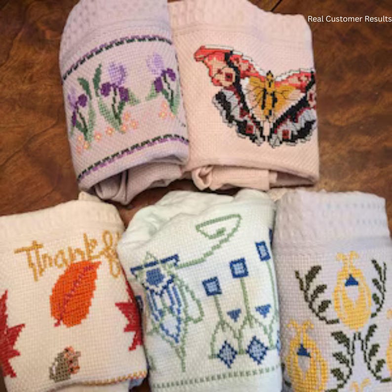 May include: Five folded fabric towels featuring cross-stitch embroidery. Designs include purple irises, a red and yellow butterfly, an orange leaf with the text "Thankful", a green geometric floral pattern, and yellow tulip-like flowers with green stems.