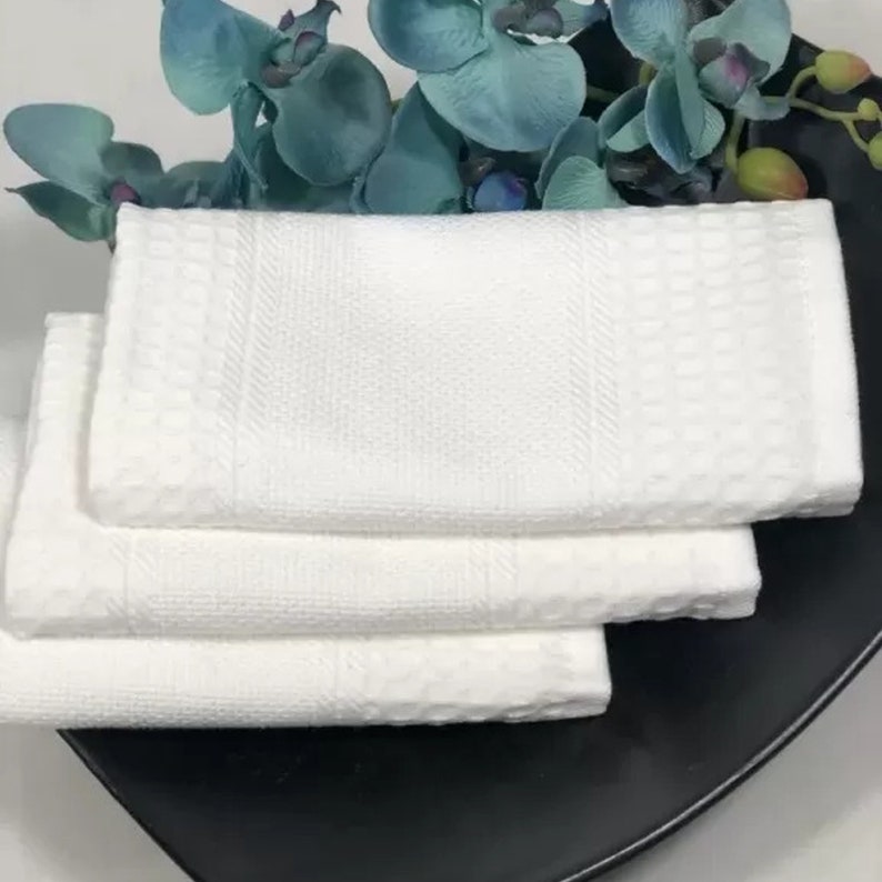 May include: Three white hand towels folded on top of each other. The towels have a waffle texture and a white border.
