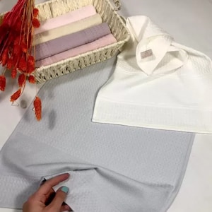May include: A light gray textured towel, a white towel with a small tag labeled SARRA, and a woven basket containing folded towels in shades of pink, beige, and mauve, accented by a cluster of dried orange-red floral stems.