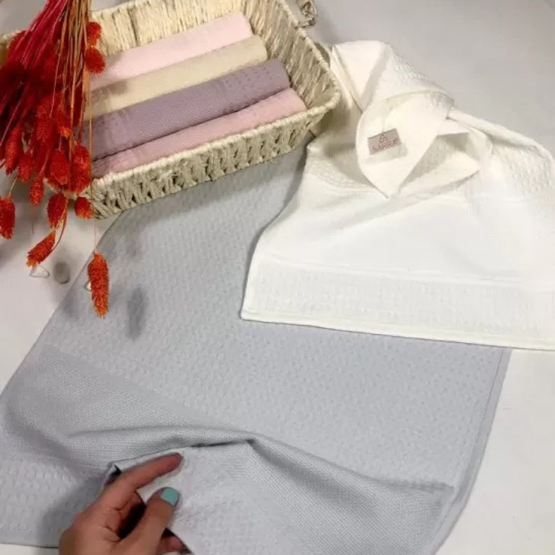 May include: A light gray textured towel, a white towel with a small tag labeled SARRA, and a woven basket containing folded towels in shades of pink, beige, and mauve, accented by a cluster of dried orange-red floral stems.